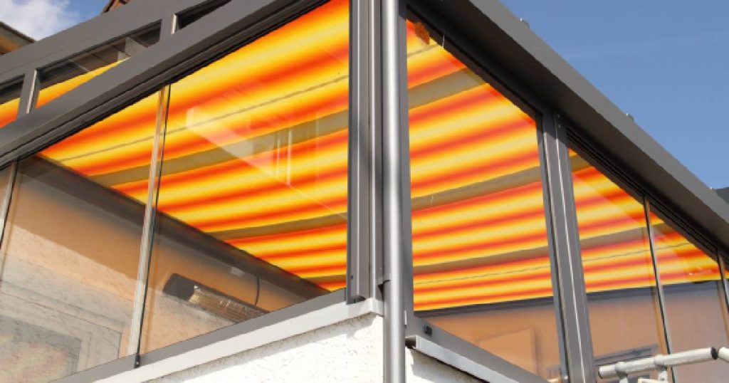 Onderaanzicht van een glazen veranda waarboven een buitenzonwering met screens gespannen is. De screen is voorzien van een oranje met gele print.