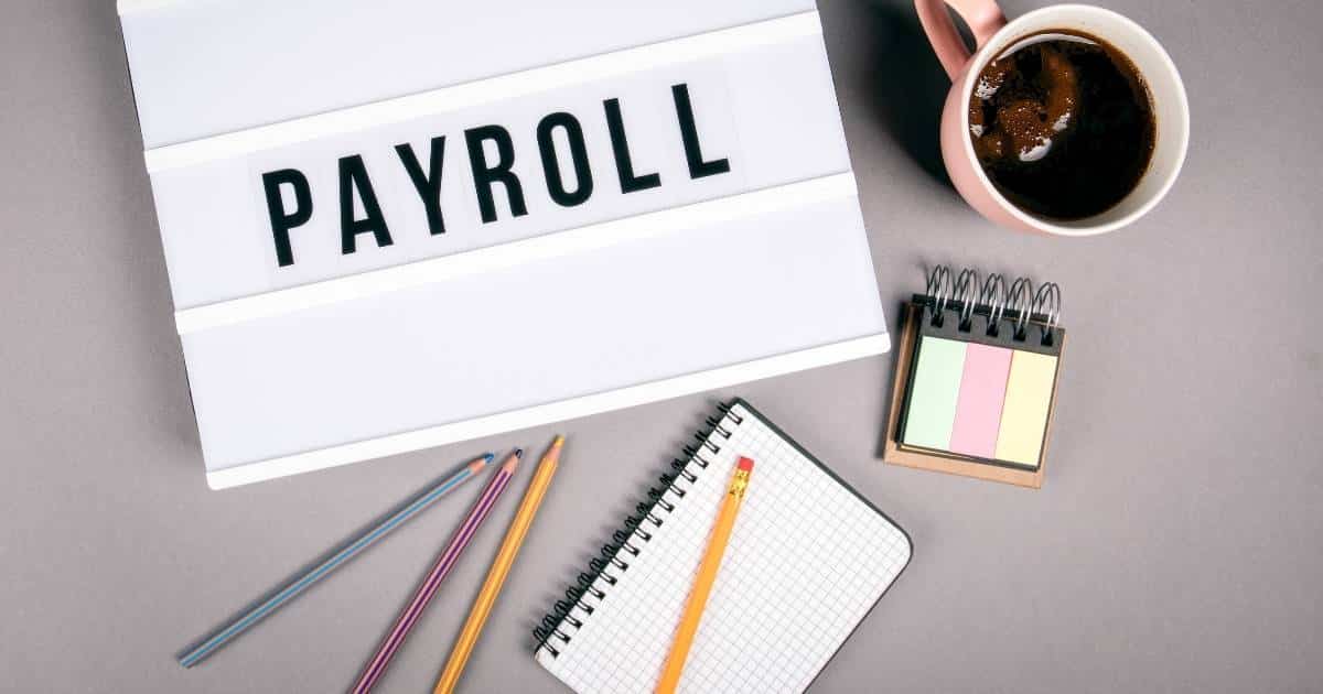Een wit bord met 3 rijen. In de middelste rij staat het woord 'payroll' in grote zwarte hoofdletters. Rechts van het bord staat een roze kop met koffie. Daaronder ligt een notitieboek met groene, roze en gele post-its. Daaronder ligt een geruit notitieblok waarop een geel potlood rust. Naast dat notiteblok liggen 3 potloden: een gele, roze en groengrijze.
