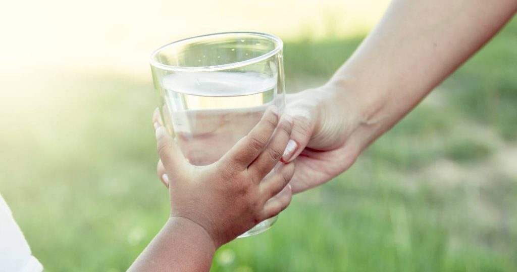 Twee handen rond een glas onthard water: een hand van een jong kindje, een hand van een volwassen vrouw. Op de achtergrond een vervaagd grasveld. 