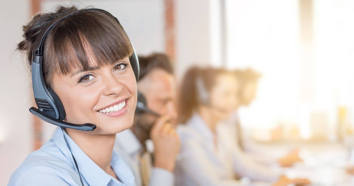 Een dame in lichtblauw hemd en met een blauwgrijze headset. Op de achtergrond zijn andere callcenter agents aan het werk.