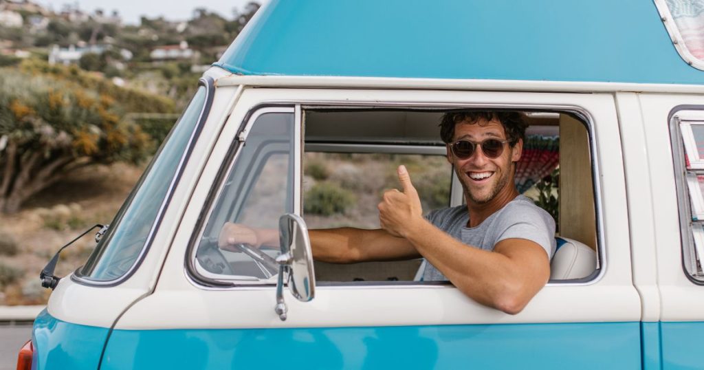 Een man met zonnebril zit achter het stuur van zijn blauw met witte bestelwagen voor particulier gebruik.
