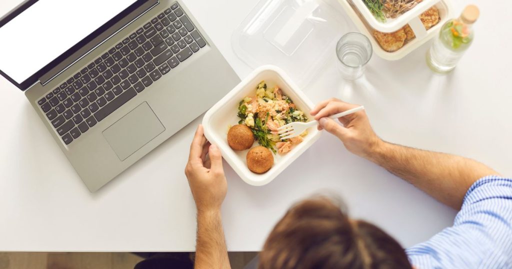Een werknemer eet lunch achter zijn laptop tijdens het werk. 
