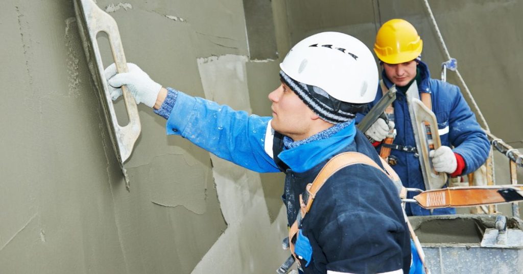 Twee vakspecialisten brengen olijfgroene of donkergroene cementpleister aan op een grijze muur. De ene draagt een witte veiligheidshelm, blauwe jas en witte werkhandschoenen. De andere draagt een gele veiligheidshelm, blauwe jas boven een rode trui, en witte werkhandschoenen. Ze hebben beide een pleistertroffel in de hand. De voorste gevelspecialist gebruikt de troffel om de groene cementpleister glad te strijken. Ze staan samen op een stelling voor de gevel.