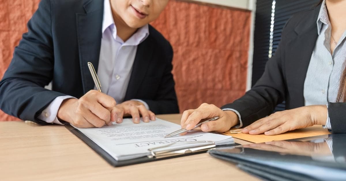 Een nieuwe werknemer in maatpak ondertekent zijn contract. Zijn nieuwe werkgever (ook in maatpak, met gestreept hem) duidt iets aan op het contract. Het contract zit in een klembord.