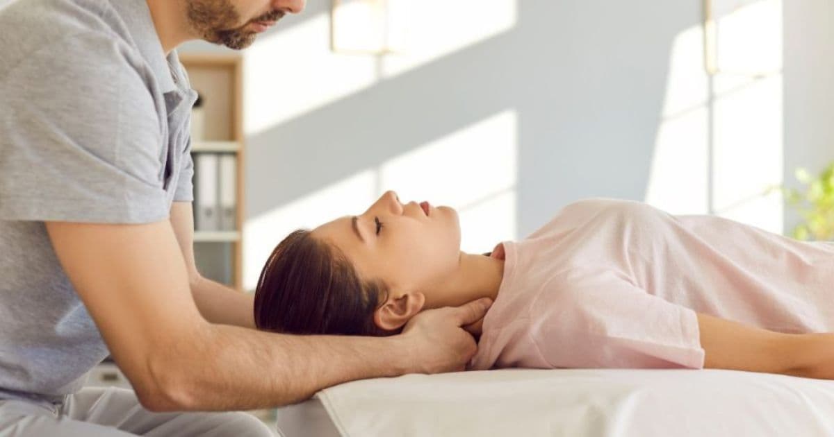 Een mannelijke chiropractor legt zijn handen in de hals van een vrouwelijke patiënt die op haar rug op een behandeltafel ligt. Haar ogen zijn gesloten. Op de muur is de schaduw van de ramen te zien.