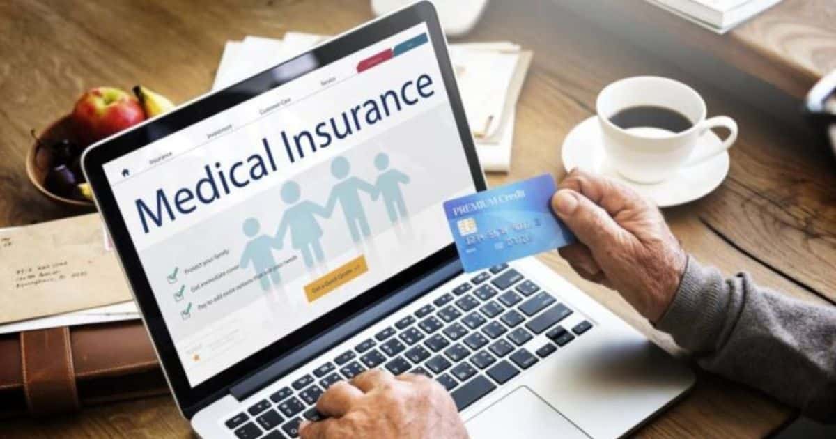 Een iets oudere hand houdt een bank- of verzekeringskaart vast voor een laptopscherm. Op het scherm staat: 'Medical Insurance'. Op de tafel staat een tas koffie en liggen enveloppen en papieren.