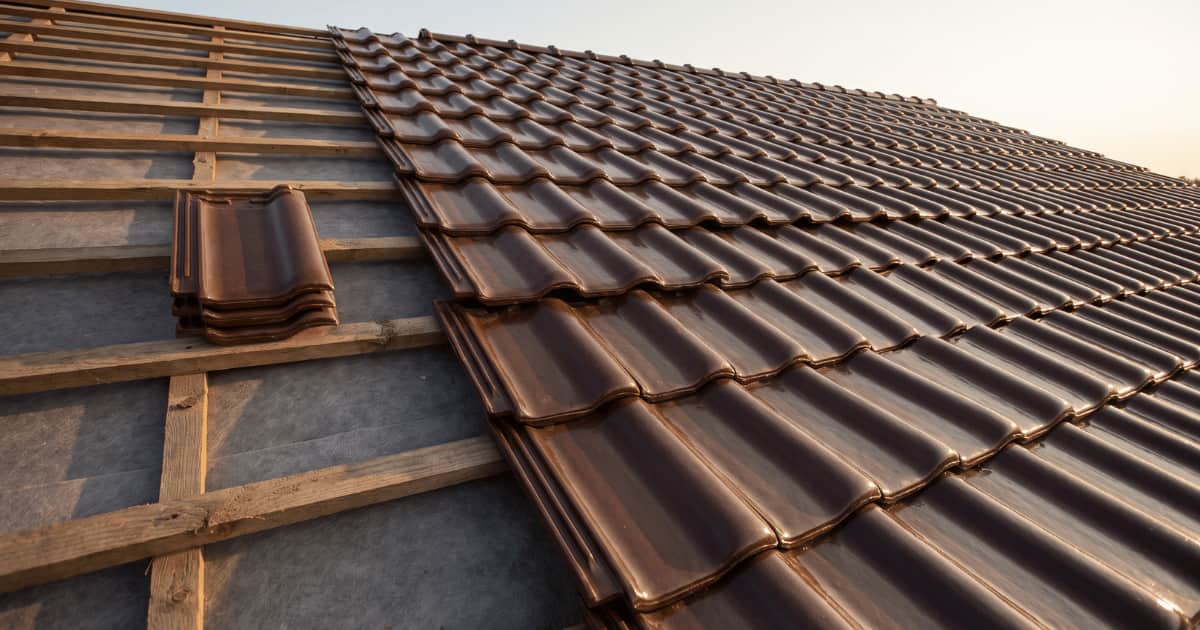 Close-up van een hellend dak in aanbouw waar rechts glanzende donkerbruine keramische dakpannen in rijen zijn gelegd en links de houten panlatten met grijs onderdak nog zichtbaar zijn, met een klein stapeltje losse dakpannen klaarliggend op de latten in warm avondlicht.