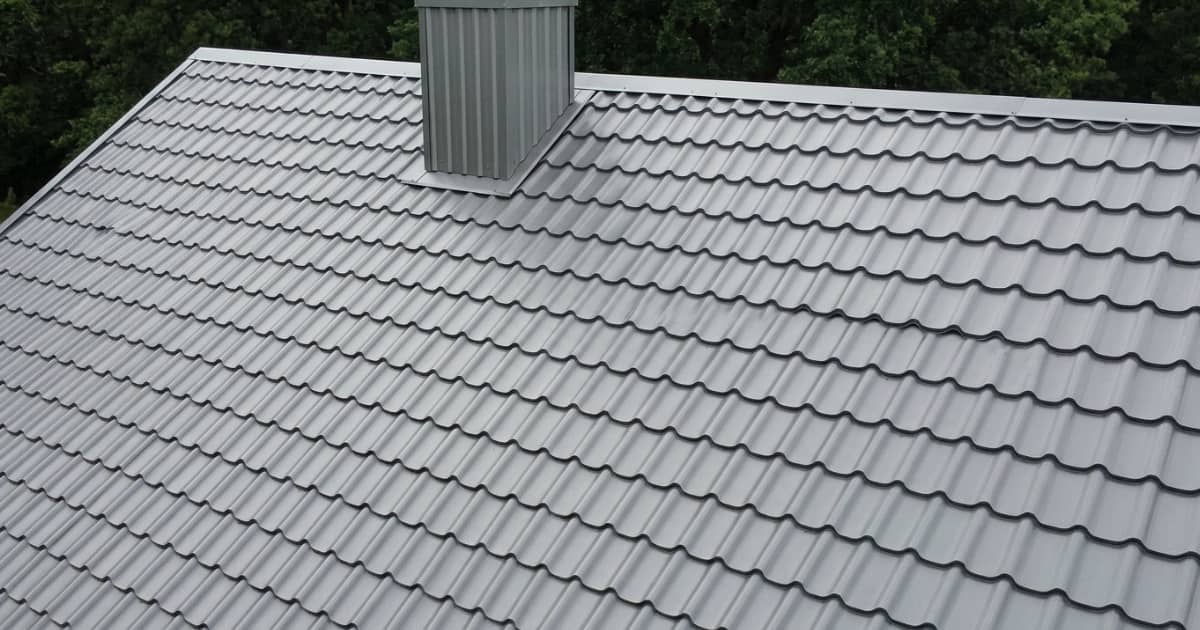 Een steil dak volledig bedekt met grijze sandwichpanelen in dakpanprofiel toont het regelmatige golvende patroon dat traditionele dakpannen nabootst met een rechthoekige grijze metalen schoorsteenbehuizing op het dakvlak en groene boomkruinen op de achtergrond.