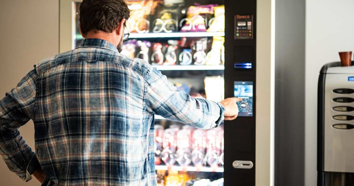 Een man met bril, baard en geruit hemd maakt gebruik van het Coca-Cola-automaat op zijn werk. Hij duwt op het bedieningspaneel om een drankje te kiezen. Naast de drankenautomaat staat een koffiemachine.