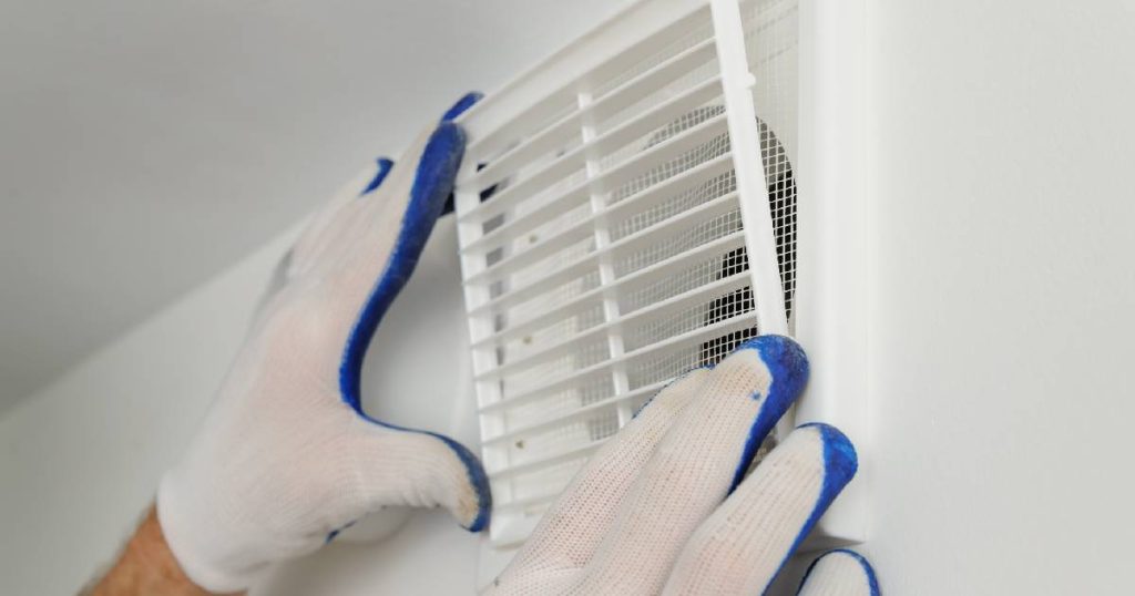 De handen van een verandaspecialist in wit met blauwe werkhandschoenen plaatsen een ventilatierooster in een veranda om condens te voorkomen.