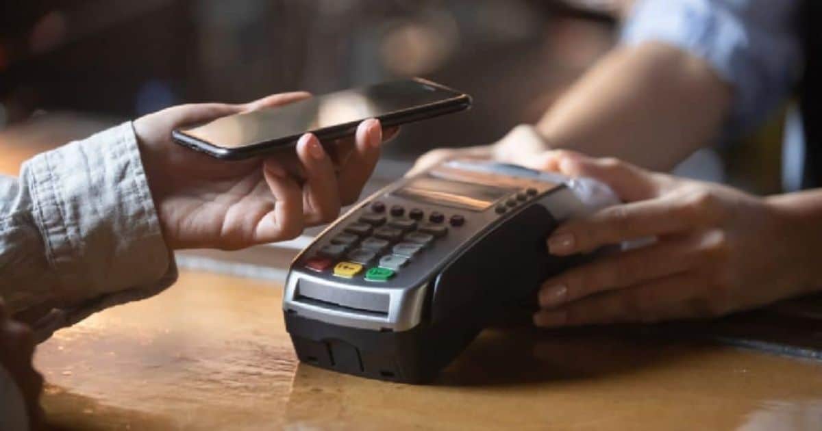 Klant houdt een smartphone boven een betaalterminal op een houten toonbank om contactloos af te rekenen, terwijl de kassamedewerker het toestel vasthoudt.