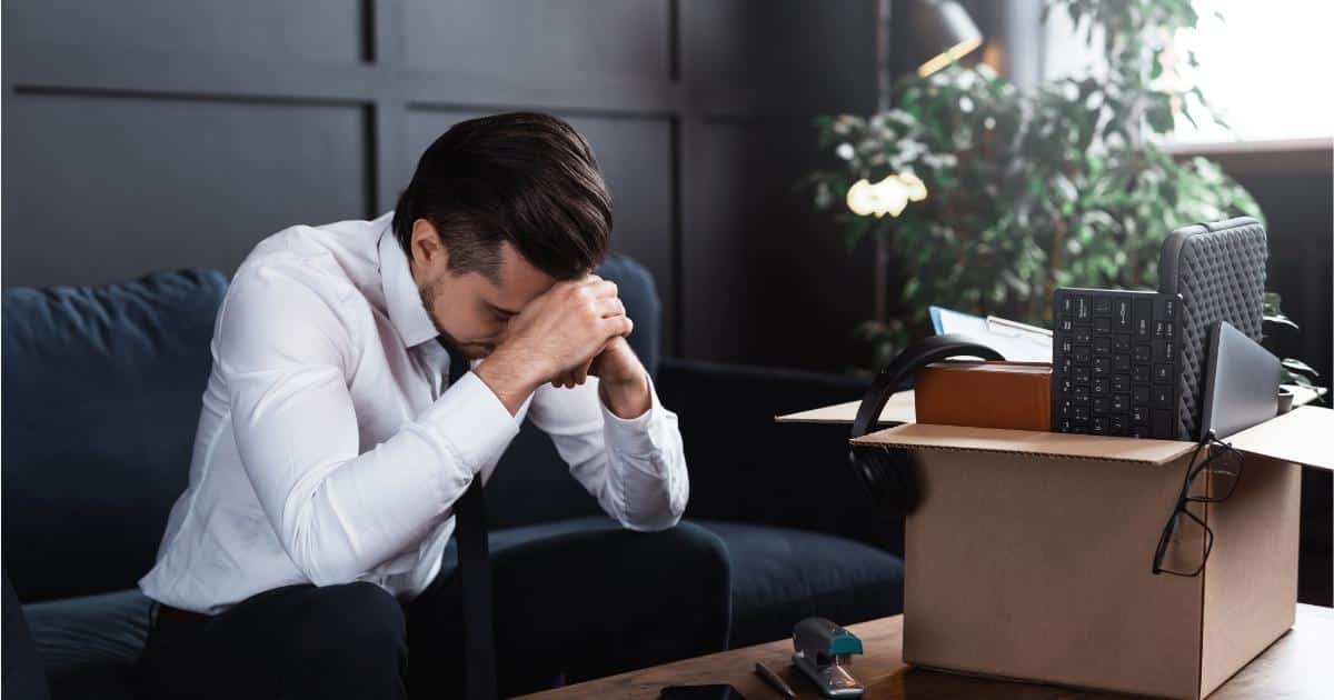 Een man met kort bruin haar en bakkebaarden, een wit hemd met zwarte das en een zwarte broek, zit teneergeslagen op een zwarte zetel voor een houten salontafel. Hij leunt met zijn hoofd tegen zijn over elkaar geslagen handen. Op de tafel voor hem staat een kartonnen doos met werkgerei: een toetsenbord, laptophoes, koptelefoon, klembord, zwarte bril en boeken. Naast de doos staat een blauw nietjesmachine.