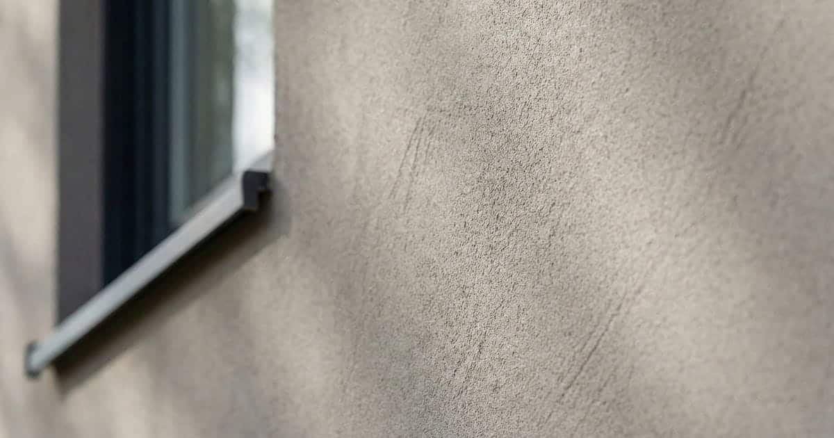Close-up van een grijze crepi gevel met zichtbare textuur en een raamkozijn op de achtergrond
