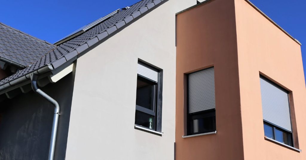 Een egale, witte crepi gevel met uitstekend gedeelte dat bekleed werd met strakke, zalmroze crepi. De woning heeft een donkergrijs pannendak, en drie strakke ramen.