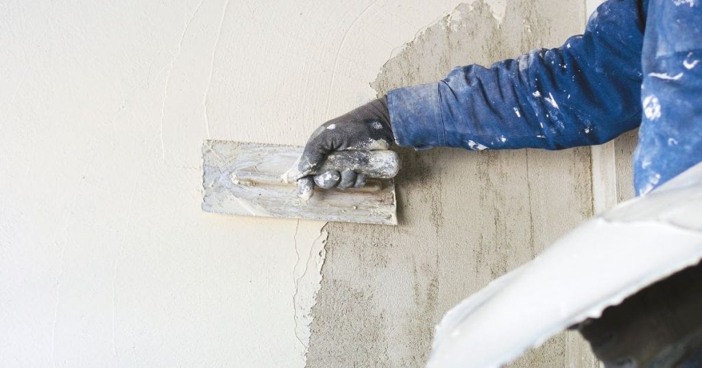 Een gevelspecialist gebruikt een pleistertroffel om dikke, witte minerale crepi aan te brengen op een grijze gevel. Hij draagt een blauwe werktrui en zwarte werkhandschoenen. Zijn kleding is besmeurd met beetjes crepi.