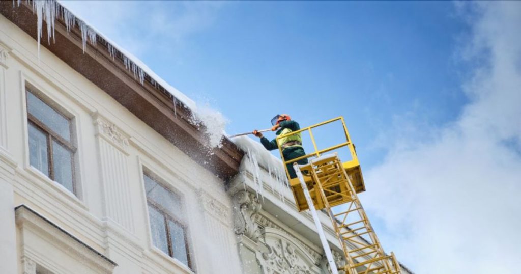 Een werkman of -vrouw is het dak en de bijhorende dakgoot vanop een gele steiger of hoogtewerker onder hogedruk aan het afspuiten.