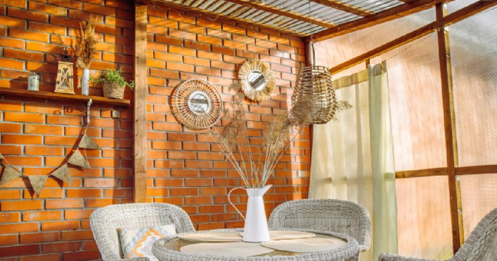 Een veranda tegen een bakstenen muur met dakbedekking en wanden uit polycarbonaat. In de veranda staan een tafel met vaas en drie stoelen. Tegen de bakstenen muur hangt een plankje met plantjes en kaarshouders.