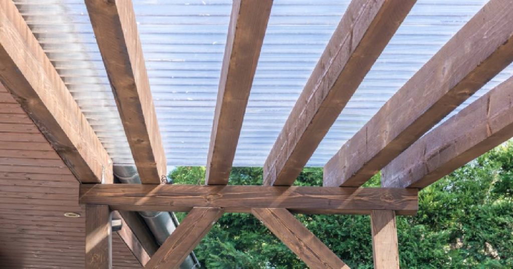 Een veranda met dakbedekking uit polycarbonaat op een houten frame. Achter de veranda is een struik zichtbaar. Over het verandadak loopt een dakgoot.