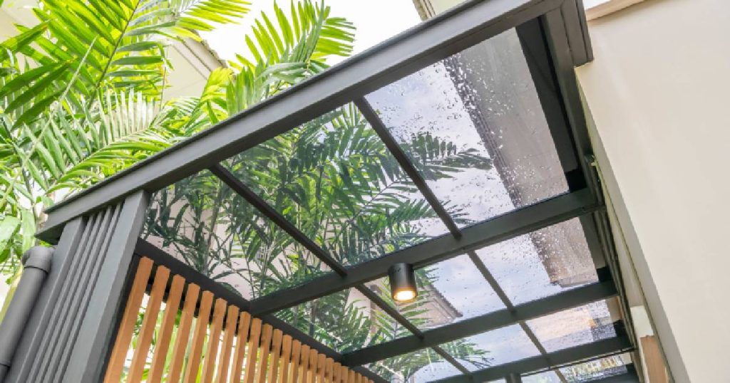 Een veranda met dakbedekking uit polycarbonaat, een alternatief op glas met een lagere prijs. In deze uitvoering met platte kanaalplaten op een zwart, aluminium frame. Door het transparante dak zijn palmbladeren en een blauwe lucht zichtbaar.