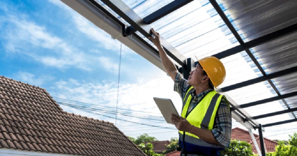 Een verandaspecialist inspecteert een veranda met polycarbonaat dakbedekking. Hij draagt een fluohesje over een geruit hemd en een gele veiligheidshelm.