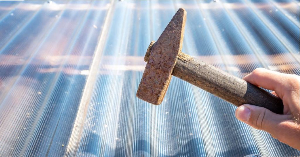 Een veranda met dakbedekking uit polycarbonaat golfplaten. Een man houdt een roestige hamer boven de golfplaten om de stevigheid en slagvastheid van het materiaal te demonstreren.
