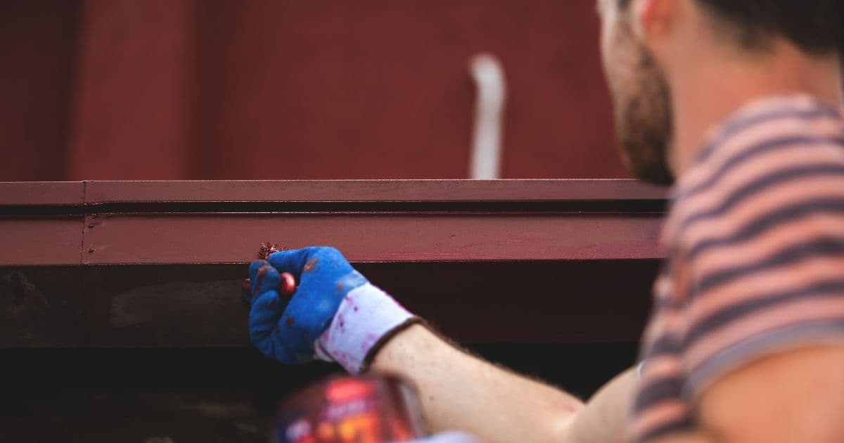 Een man in streepjes t-shirt wrijft met zijn verfborstel over de onderkant van een rechthoekige dakgoot. Hij draagt daarbij blauwe werkhandschoenen om te vermijden dat zijn handen vol verfspatten zitten.