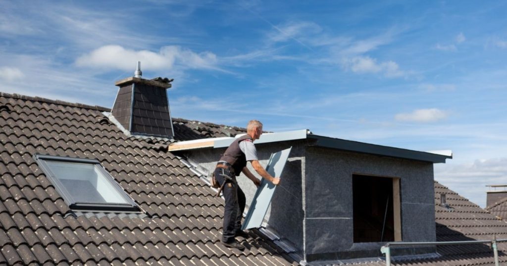 Een man in grijze overall is bezig met het plaatsen van een isolatie en afwerkingsplaat tegen de zijwand van een dakkapel.