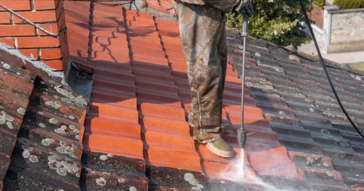 Een man is een dak aan het reinigen met een hogedrukspuit. De helft van het dak is schoon, de andere helft is nog bedekt met mos.