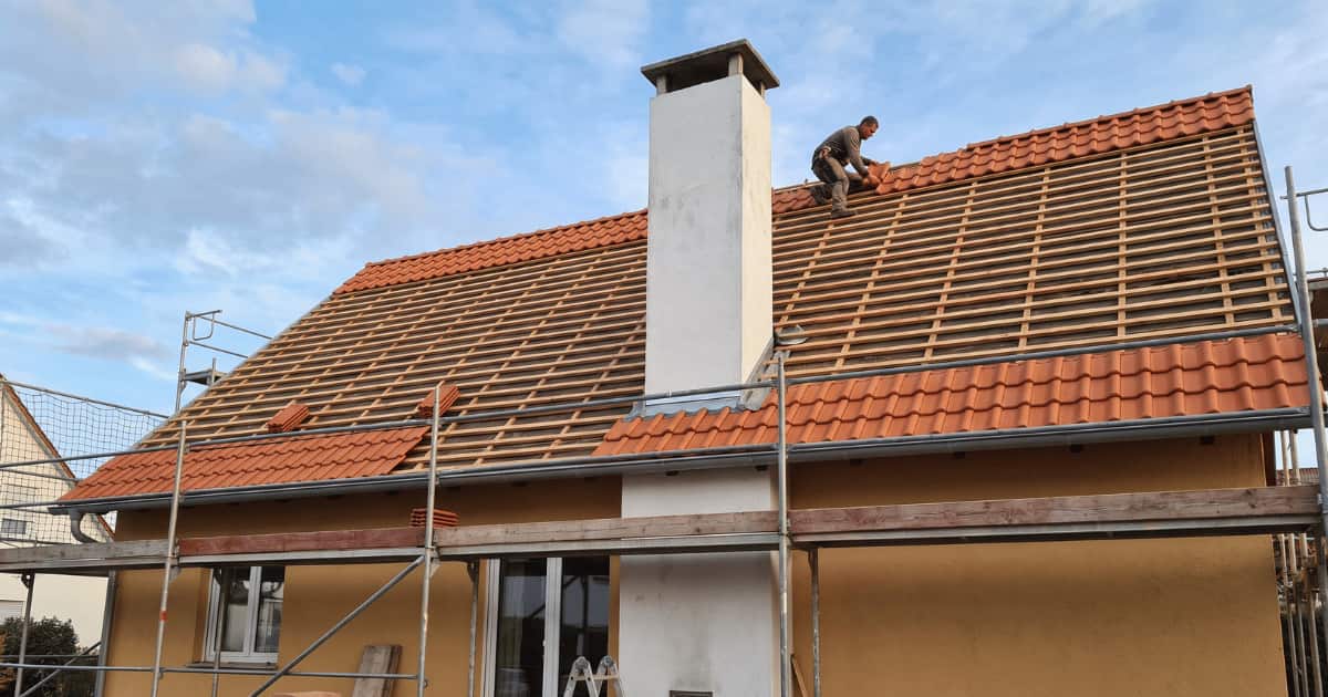 Dakwerker hurkt nabij de nok van een hellend dak en plaatst oranjerode keramische dakpannen op blootliggende houten panlatten terwijl de linkerzijde van het dak al volledig bedekt is en rondom het huis metalen stellingen staan opgesteld langs een okergele gevel met een grote witte schoorsteen.