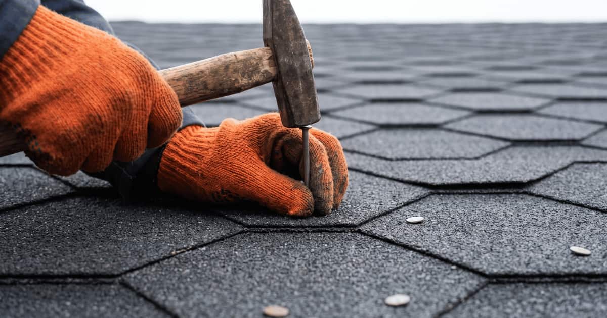 Twee handen in oranje werkhandschoenen slaan met een versleten houten hamer een nagel in donkergrijze zeshoekige bitumen dakshingles terwijl het overlappende patroon van de shingles zich onscherp naar de achtergrond uitstrekt.