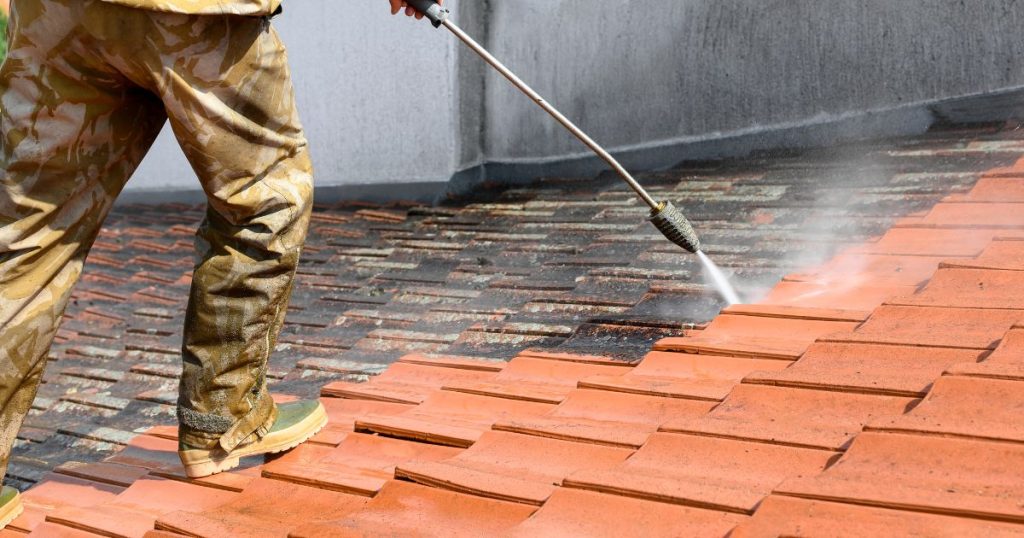 Een dakwerker gebruikt een hogedrukreiniging om sterk verontreinigde oranje dakpannen te reinigen. Rechts in beeld zijn de vuile, bijna zwart besmeurde dakpannen; links de gereinigde oranje dakpannen.