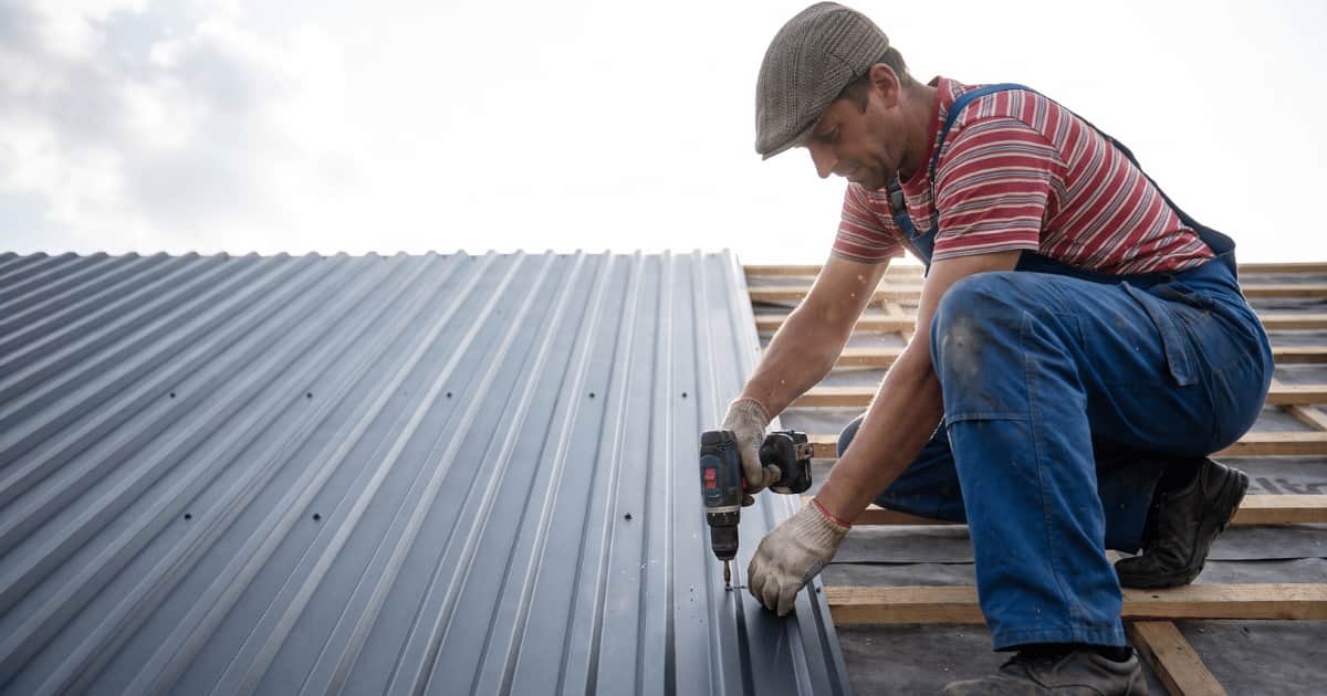 Een dakwerker in blauwe overall en pet hurkt op houten daklatten en schroeft met een accuboormachine een grijze metalen golfplaat vast terwijl de verticale ribben van de plaat omhoog lopen tegen een heldere lucht met wolken.