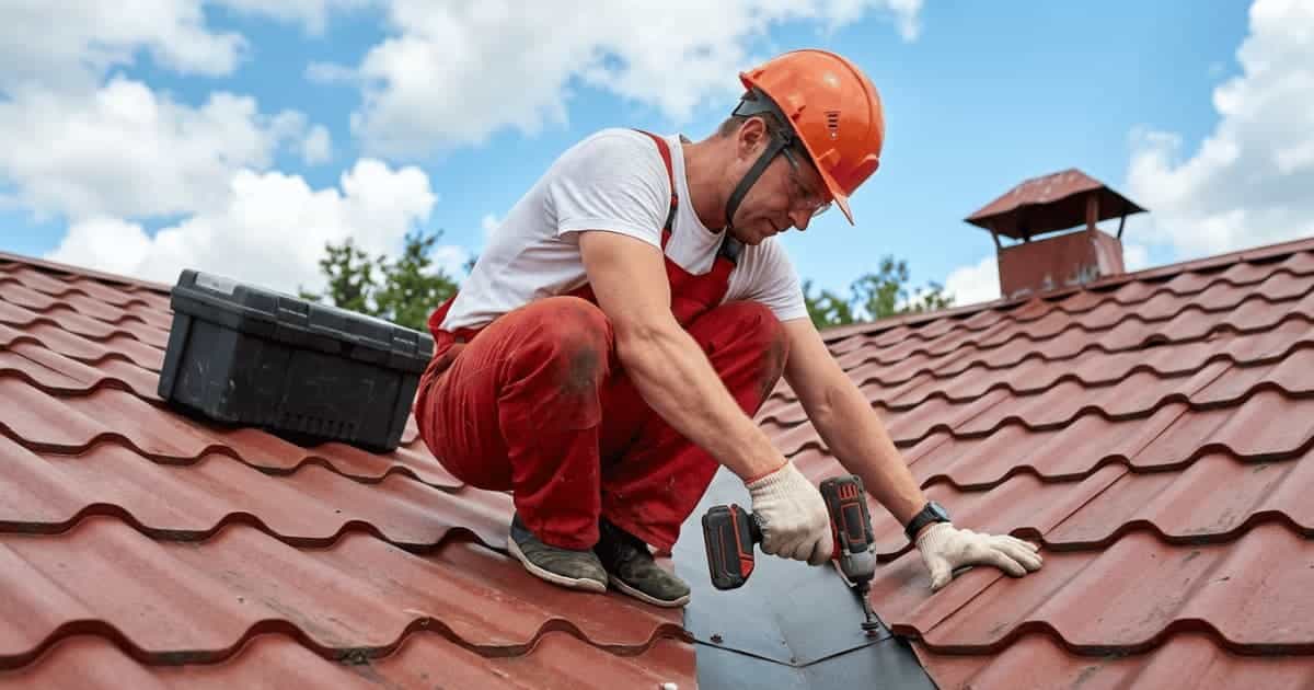 Een dakwerker in wit shirt rode overall en oranje helm hurkt op een hellend dak met roodbruine metalen dakpanplaten en schroeft een donkere afwerkingsplaat vast met een accuboormachine terwijl een zwarte gereedschapskoffer naast hem op het dak staat.