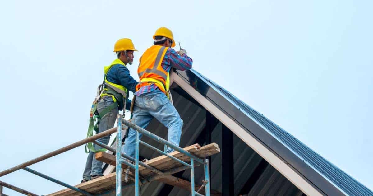 2 Dakwerkers met fluohesje, gele helm en veiligheidsriemen werken vanop een stelling aan de daknok van een aluminium dak.