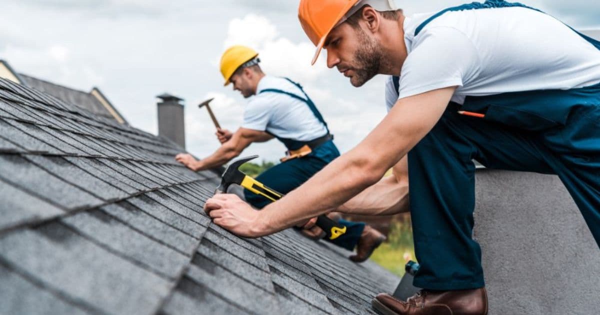 2 dakwerkers in blauwe overall en met een veiligheidshelm (geel en oranje) zijn druk in de weer met het vastnagelen van de donkere leien.