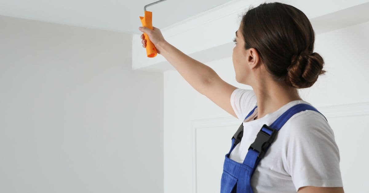 Een dame in blauwe overall schildert een plafond wit met een verfroller met oranje handgreep.