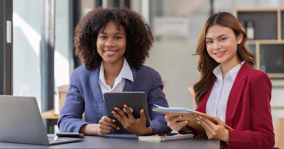 Twee accountants of boekhouders zitten naast elkaar aan een grijze tafel. De linkse heeft bruin krullend haar en draagt een blauw vest boven een wit hemd. Ze heeft een tablet in haar handen en glimlacht. Voor haar op de grijze tafel staat een laptop. De rechtse heeft bruinblond, golvend haar en heeft een dossier in handen. Ze draagt een rood vest boven een wit hemd en glimlacht. Onder het dossier in haar handen ligt een notitieblok met ringen, met daarop een pen.
