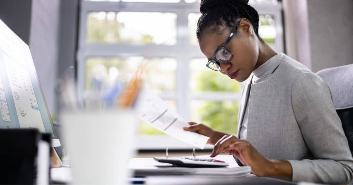 Een boekhouder draagt een grijs mantelpak en een bril met zwart montuur. Ze tikt, met haar linkerhand, iets in op een rekenmachine. In haar rechterhand houdt ze een document of factuur vast. Op de voorgrond, vervaagd, een houder met pennen. Voor de boekhouder staat een computerscherm. Ze zit op een zwarte bureaustoel.