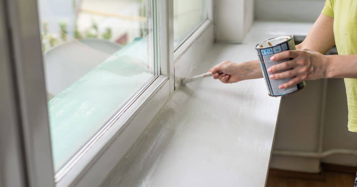 Een dame houdt in haar linkerhand een verfpot vast terwijl ze met een fijne borstel in de rechterhand een raamkozijn in aluminium schildert.