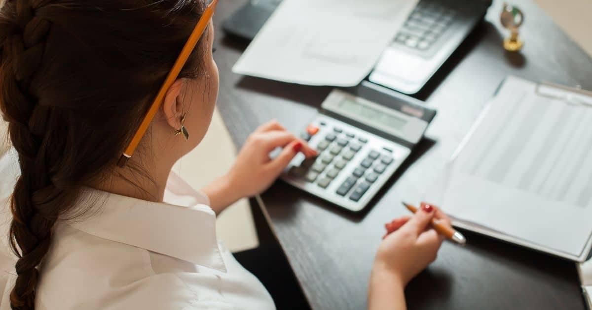 De schouder en het hoofd van een boekhouder met bruin haar in een vlecht. Ze draagt een wit hemd en heeft een oranje potlood achter haar rechteroor. In haar rechterhand houdt ze een pen vast, met de linkerhand tikt ze iets in op een rekenmachine. De rekenmachine ligt op een houten tafel. Links van de rekenmachine ligt een klembord met een lijst, boven de rekenmachine staat een laptop waarop een document ligt.