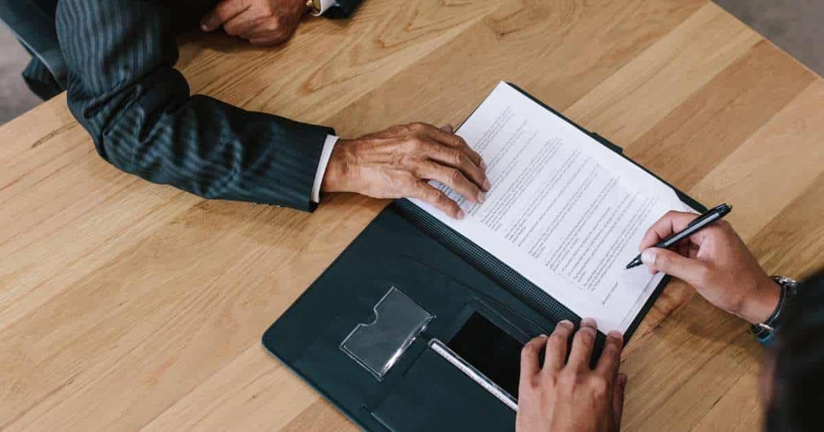Een contract in een zwarte map ligt op een lichte houten tafel. De handen van een man met horloge ondertekenen het contract met een zwarte pen. Aan de overkant van de tafel zit een tweede man, met wit hemd en zwart gestreepte blazer. Hij houdt zijn hand boven de bovenste linkerhoek van het contract.