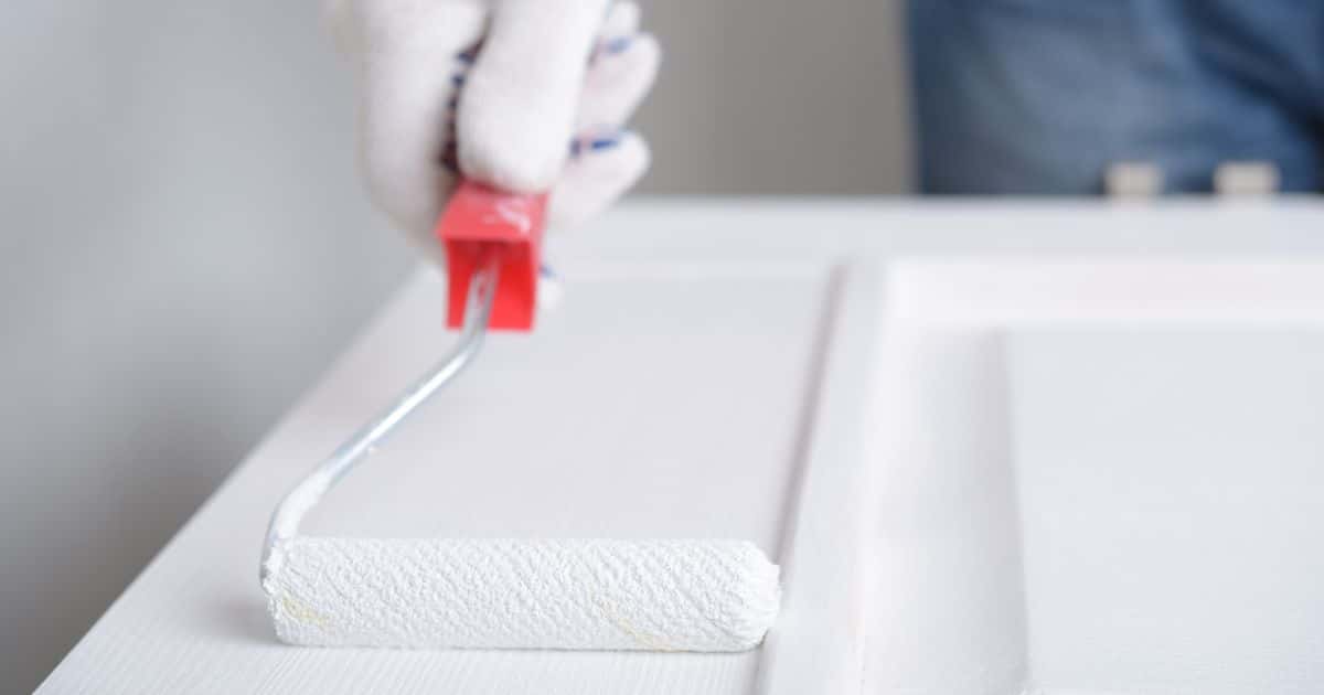Een persoon verft een deur wit met een verfroller met rood handvat. Hij of zij draagt daarbij een witte handschoen.