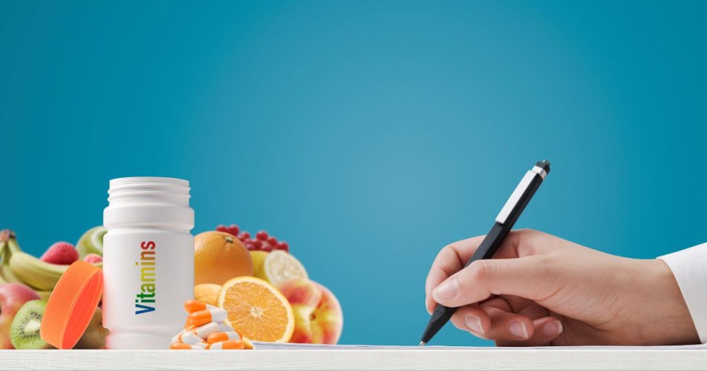 Een stapel fruit, een potje met opschrift 'vitamines' en een hand met pen dat notities maakt op een blad.