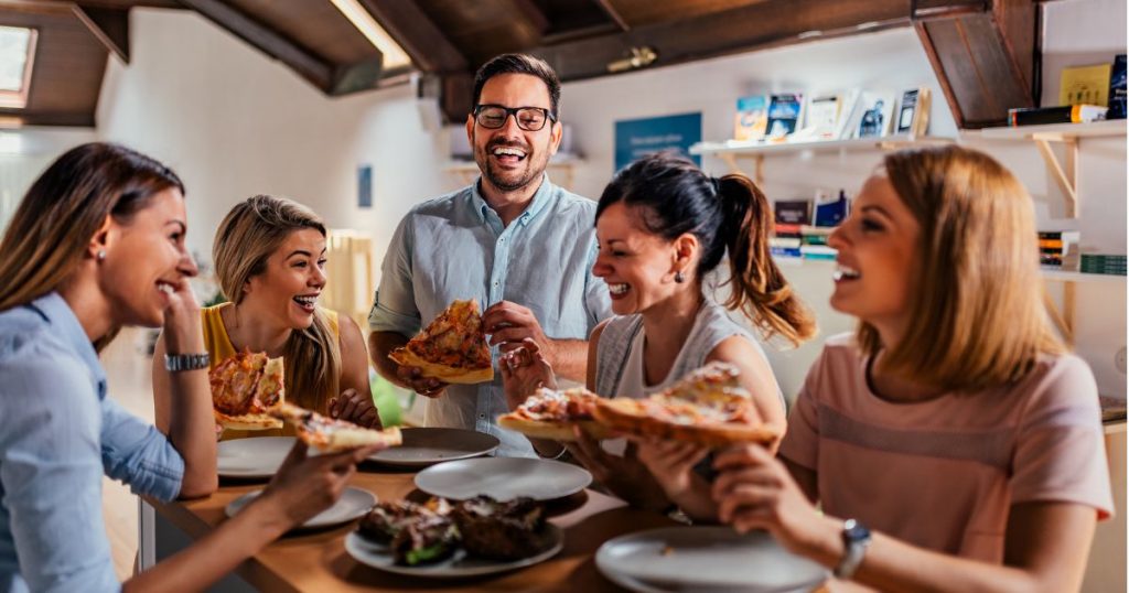 4 Dames en 1 man die samen genieten van een gezamenlijke maaltijd. Op de tafel liggen ook nog 4 lookbroodjes op een bord. 