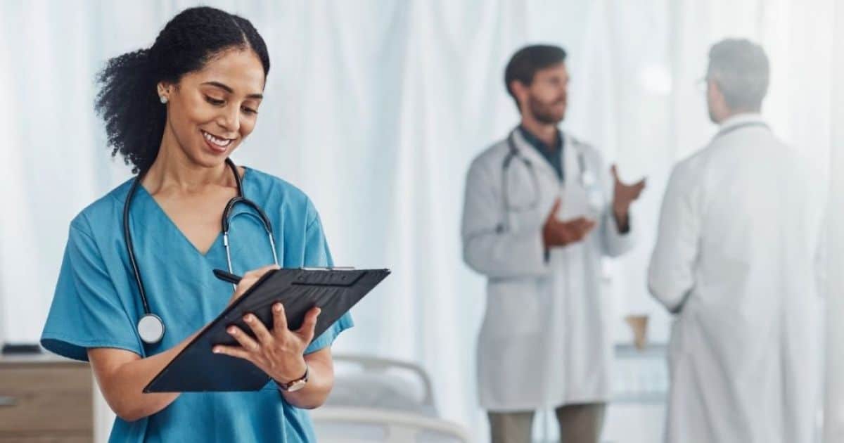 Een jonge dokter of verpleegster met een blauw tenue schrijft iets op een blad op een klembord. Ze heeft een stethoscoop rond haar nek. Op de achtergrond staan 2 mannen in dokterskledij te praten.