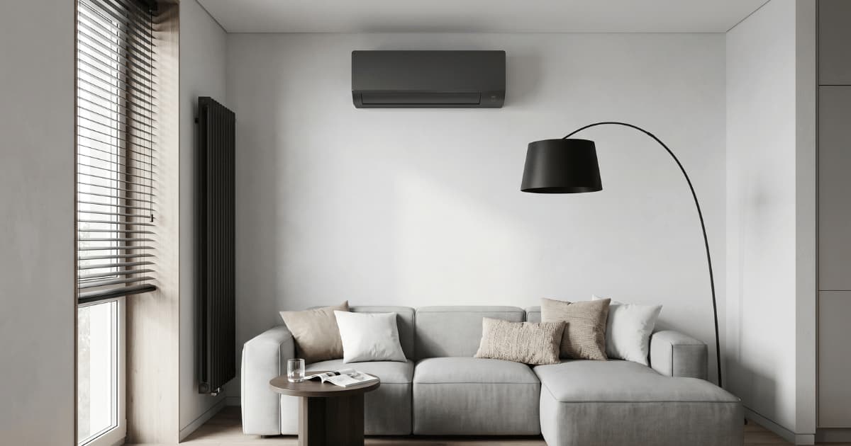 Een strakke donkergrijze airco-binnenunit hangt hoog aan de witte muur van een moderne appartementswoonkamer met een lichtgrijze zitbank, zwarte booglamp en horizontale jaloezieën voor het raam.