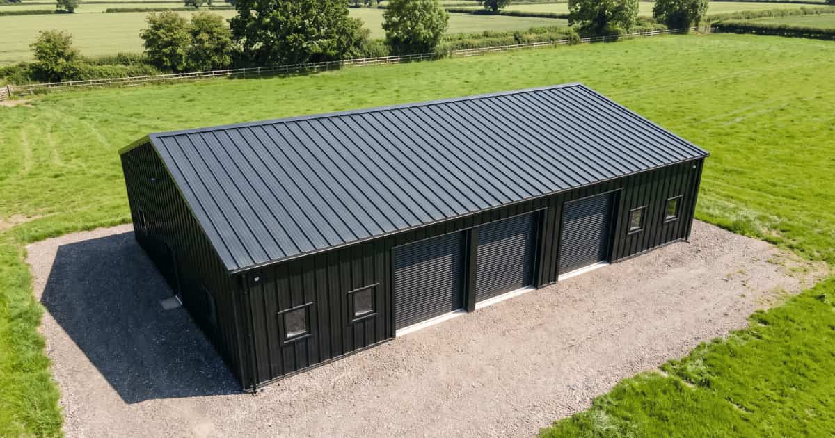 Een donkere rechthoekige loods met een zadeldak bedekt met antraciet metalen profielplaten staat op een grindverharding te midden van groene weilanden in een landelijke omgeving onder een zonnige lucht.