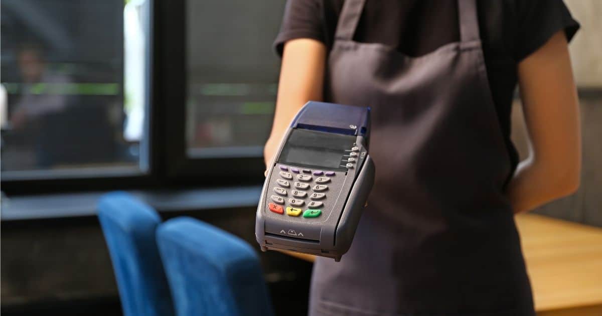 Een medewerker in een horeczaak (met zwart t-shirt en donkergrijs schort) houdt een draagbare betaalterminal voor zich.