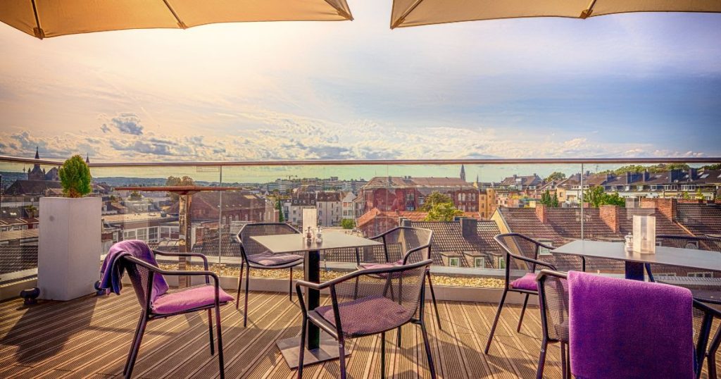 Een dakterras met 6 stoelen en 2 tafels onder twee parasols, op een houten vloer die rust op drukvaste isolatieplaten. Het terras geeft uit op de daken van lager gelegen woningen in een dorp. Op de achtergrond een lichtblauwe hemel met wolken.