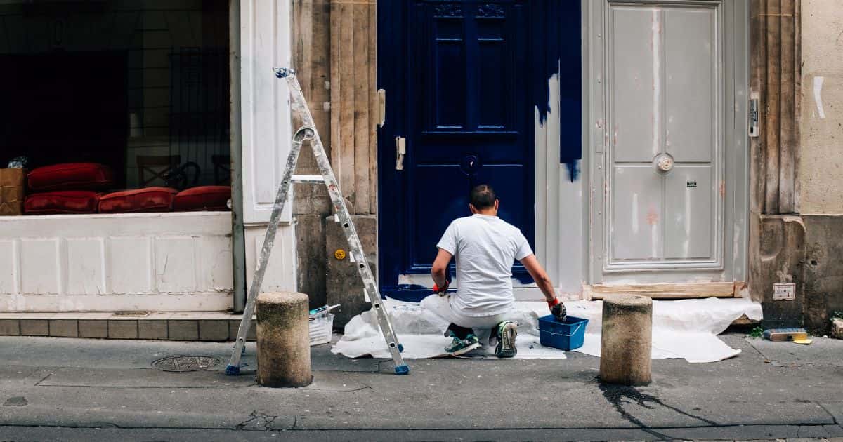 Een man staat gehurkt op een witte doek met zijn hand in een blauwe verfbak. Houdt hij een borstel of een verfroller vast? Zeker is dat hij de deur voor hem een donkerblauwe kleur geeft. Naast hem staat ook een lage ladder.